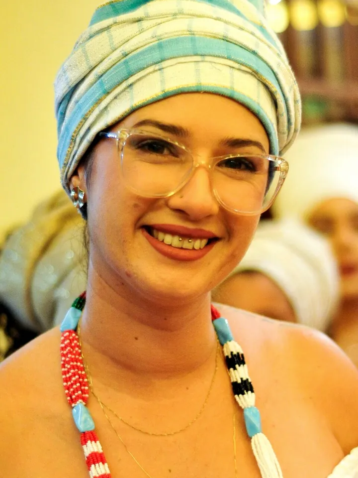 Bruna de Iemanjá sorrindo, com turbante azul e branco e guias coloridas, no terreiro