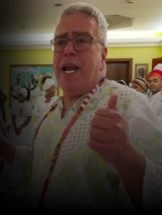 Babalorixá Pedro de Oxum falando no terreiro, de roupa branca rendada e guias, com filhos da casa ao fundo