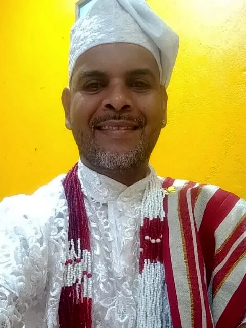 Thiago d'Xangô sorrindo, com roupa branca de renda, gorro branco e pano listrado vermelho